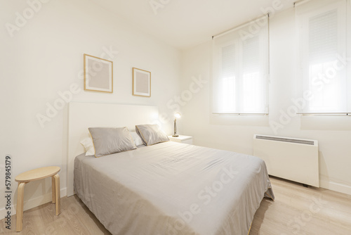 Bedroom with a double bed with wooden bedside tables  a gray bedspread with cushions  double windows with roller shutters and an electric heater