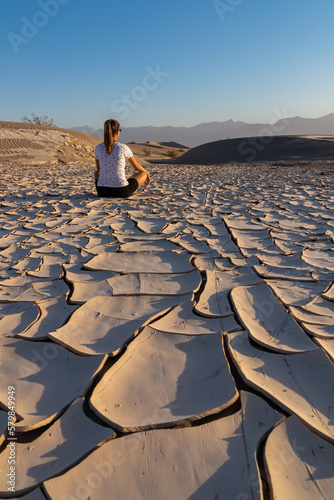 Woman sitting on dry cracked clay crust at Mesquite Flat Sand Dunes in Death Valley National Park, California, USA. Looking at pattern Mojave desert in summer with Amargosa Mountain Range in the back photo