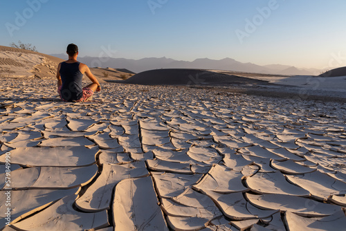 Man sitting on dry cracked clay crust at Mesquite Flat Sand Dunes in Death Valley National Park, California, USA. Looking at pattern Mojave desert in summer with Amargosa Mountain Range in the back photo