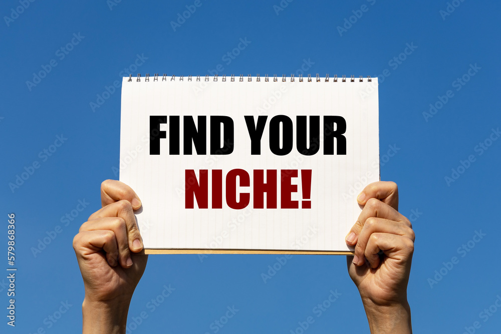 Find your niche text on notebook paper held by 2 hands with isolated blue sky background. This message can be used as business concept about finding your niche.