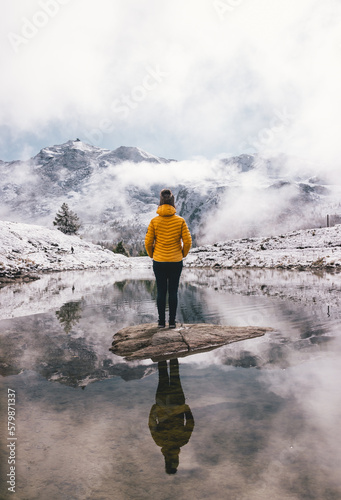person in yellow jacket reflection in a calm pond in the mountains with snowy mountains in distance