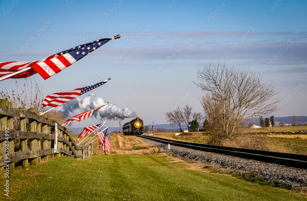 Fototapeta premium A View of a Classic Steam Passenger Train Approaching, With American Flags Attached to a Fence on a Sunny Summer Day