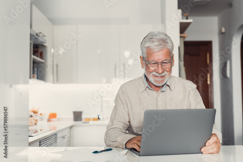 Aged remote worker. Concentrated senior male in glasses work on laptop from home office read email electronic document. Old age man employee freelancer sit at kitchen table by pc typing report online.