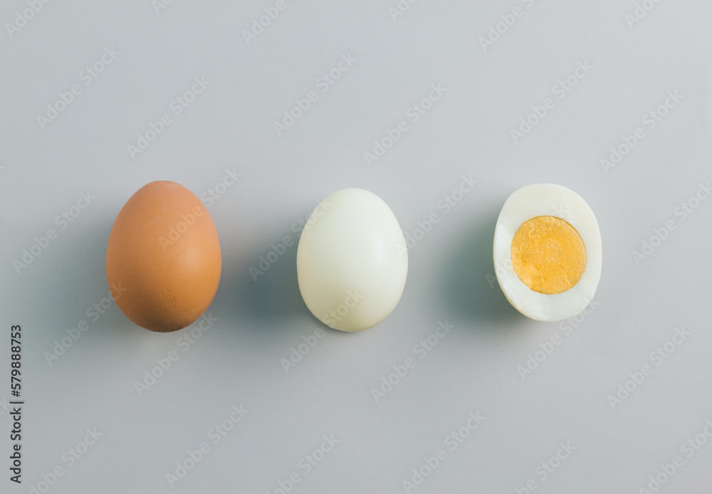 Whole chicken egg, egg without eggshell and cracked half in the middle with yolk on grey background. Minimal healthy food concept. Top view, flat lay. Whole chicken egg, egg without eggshell and cracked half in the middle with yolk on grey background. Minimal healthy food concept. Top view, flat lay.