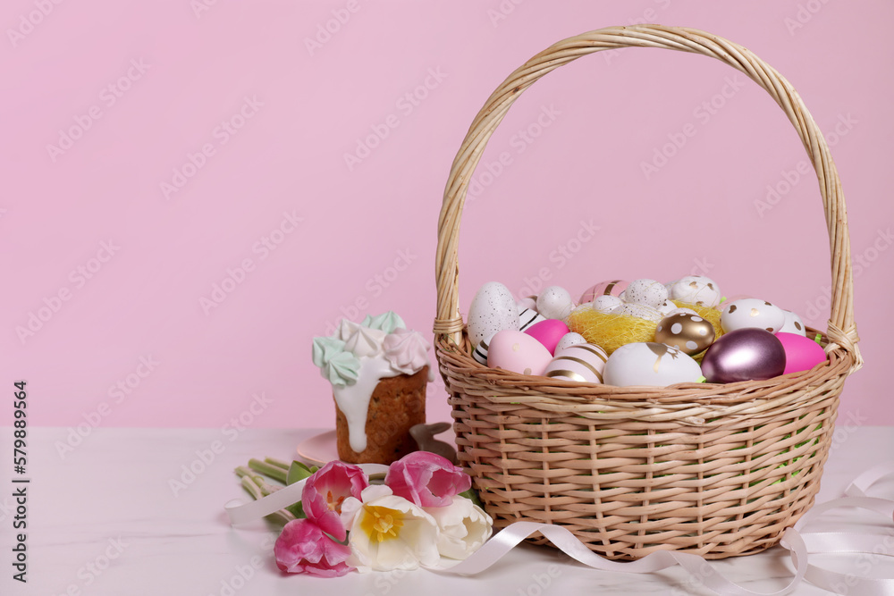 Wicker basket with festively decorated Easter eggs and beautiful tulips on white marble table against pink background. Space for text