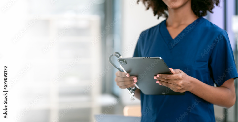 Fototapeta premium Black female nurse or doctor holding stethoscope and digital tablet standing in hospital office, copy space.