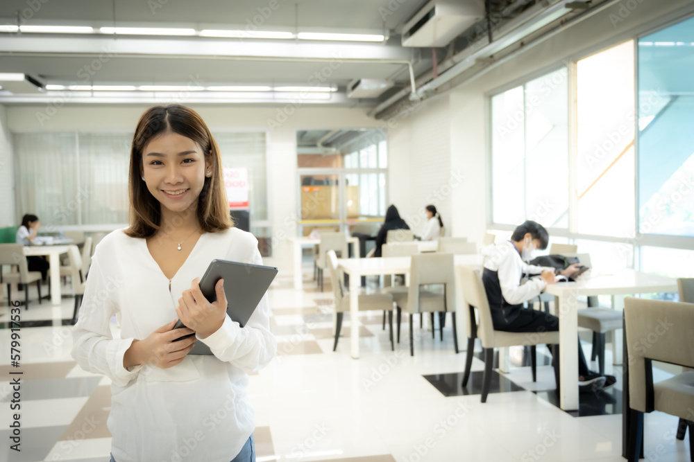 Beautiful asian female teacher is standing and looking to the camera while holding tablet with a smiling face in the library room, Copy space. Beautiful asian female teacher is standing and looking to the camera while holding tablet with a smiling face in the library room, Copy space.