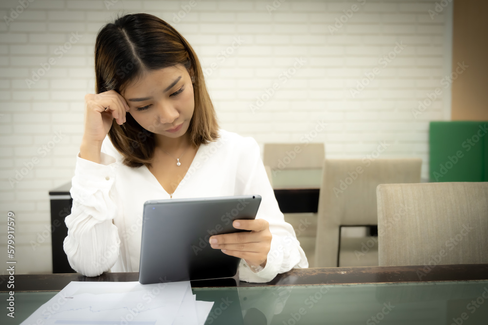 Beautiful asian female teacher sitting and looking down goes to the tablet with a serious expression on her desk. Beautiful asian female teacher sitting and looking down goes to the tablet with a serious expression on her desk.