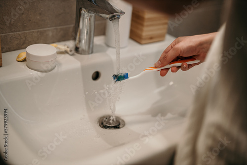 A woman is about to brush her teeth. Focus on a hand holding a toothbrush under the water in a bathroom sink.