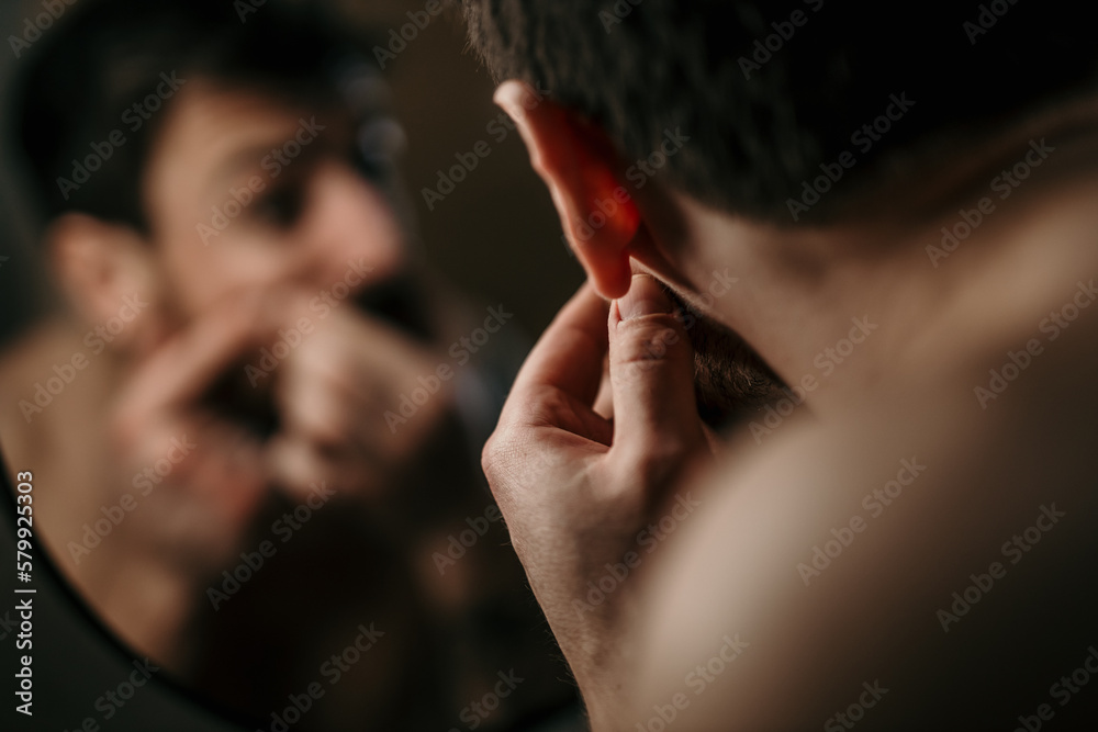 Obraz premium Shot of a young man examining his skin in the mirror at home. Health, skincare and man popping acne in face