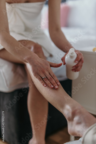 Shot of a woman sitting on a bed and using a moisturizing cream. Focus on the cream tub