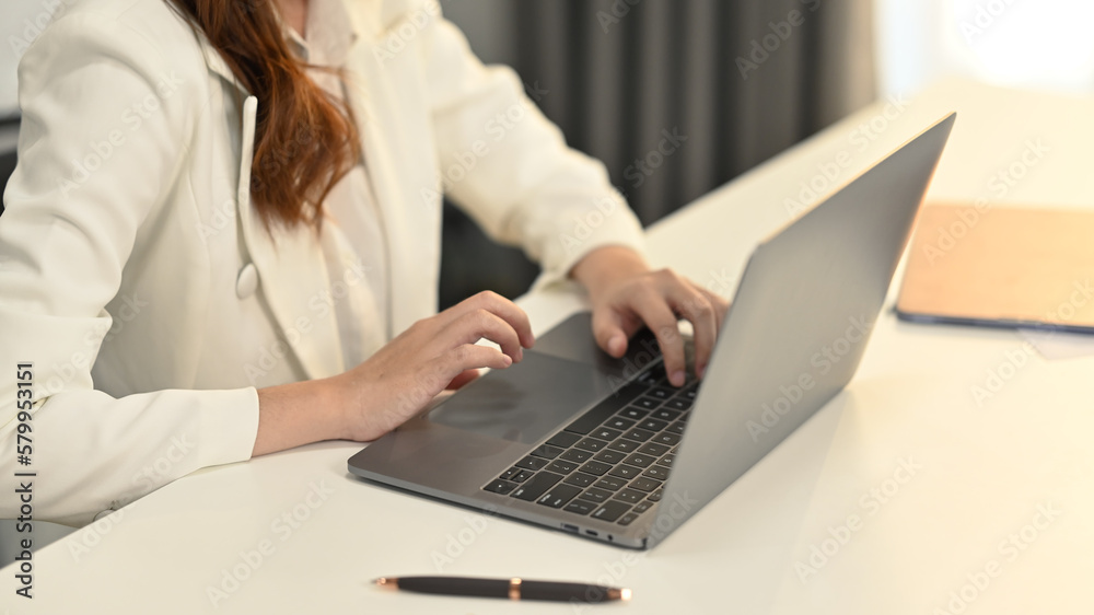 Cropped image of businesswoman in white stylish suit sitting at her working desk and using laptop computer Cropped image of businesswoman in white stylish suit sitting at her working desk and using laptop computer