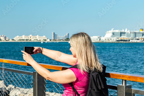 a photo of a blonde traveler girl holding a backpack while posing on a Dubai embankment with a breathtaking view of the sea and the city's modern towers