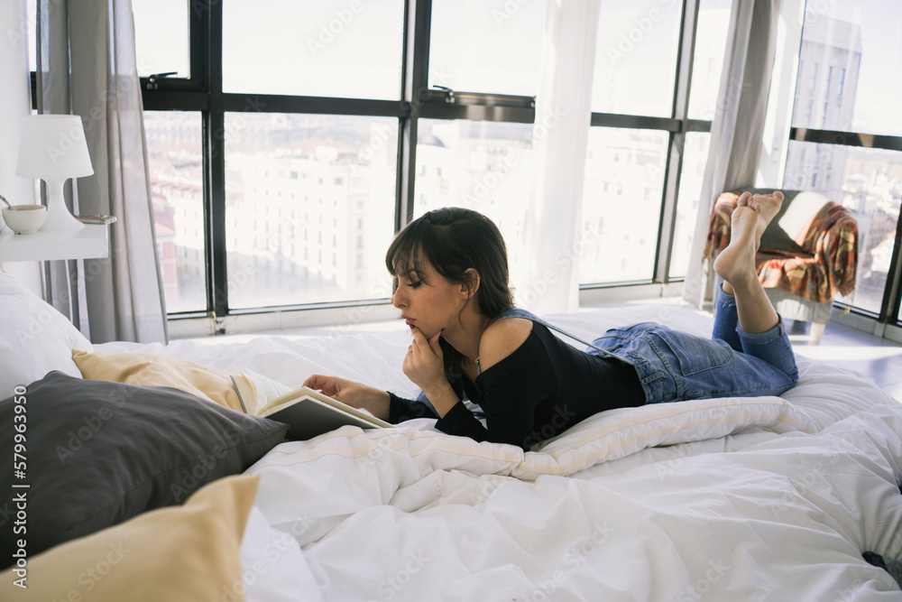 Full length of young woman reading book while lying on bed at home Full length of young woman reading book while lying on bed at home