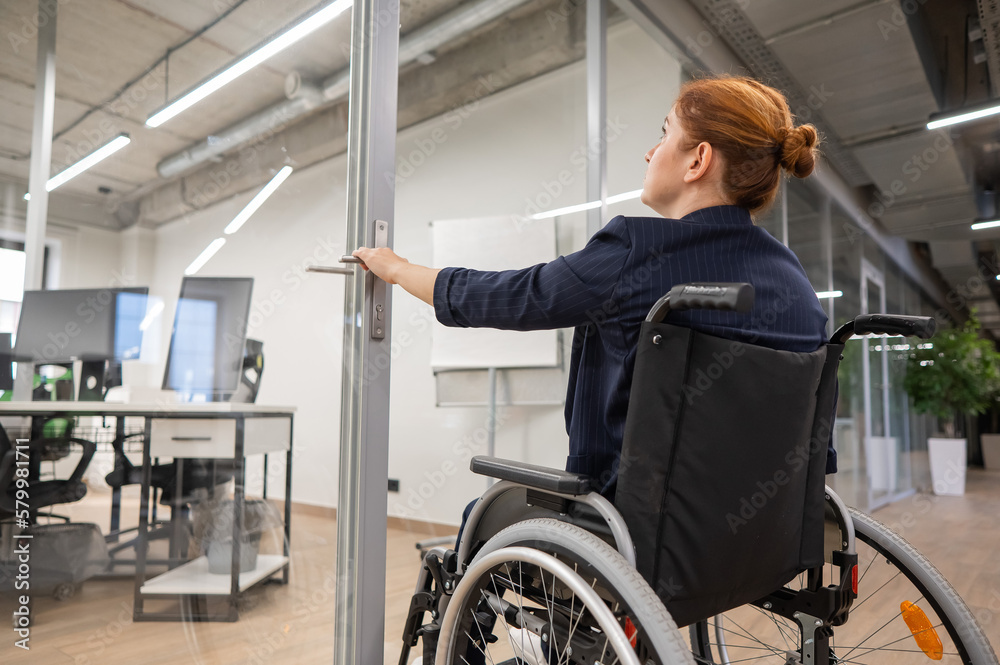 Red-haired caucasian woman in a wheelchair trying to open the door in the office. Red-haired caucasian woman in a wheelchair trying to open the door in the office.