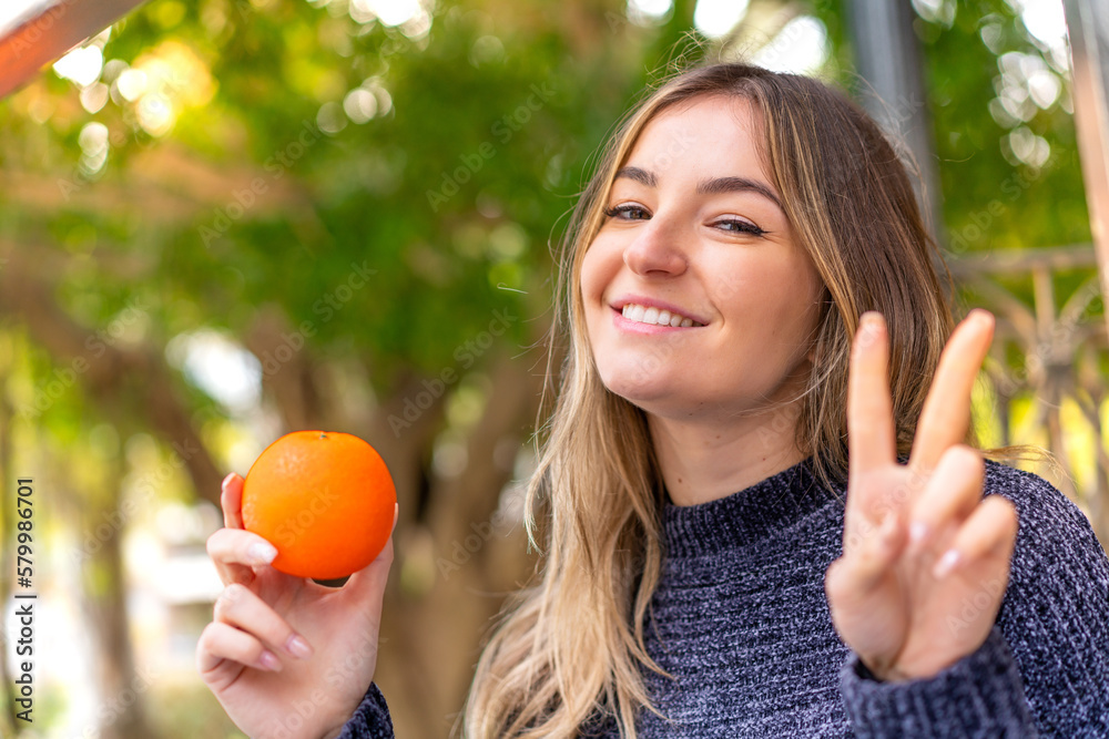 Young pretty Romanian woman holding an orange at outdoors smiling and showing victory sign Young pretty Romanian woman holding an orange at outdoors smiling and showing victory sign