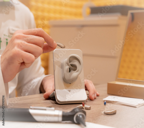 Doctor in his audiology office holding a hearing aid and in the other hand a model of a silicone ear. On his table is another hearing aid and medical devices. photo