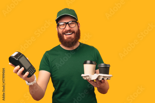Portrait of a smiling bearded man in green holding two cups and a terminal.