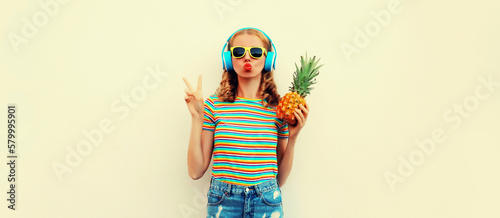 Summer portrait of cheerful happy young woman blowing her lips model posing in headphones listening to music with fresh pineapple on white background