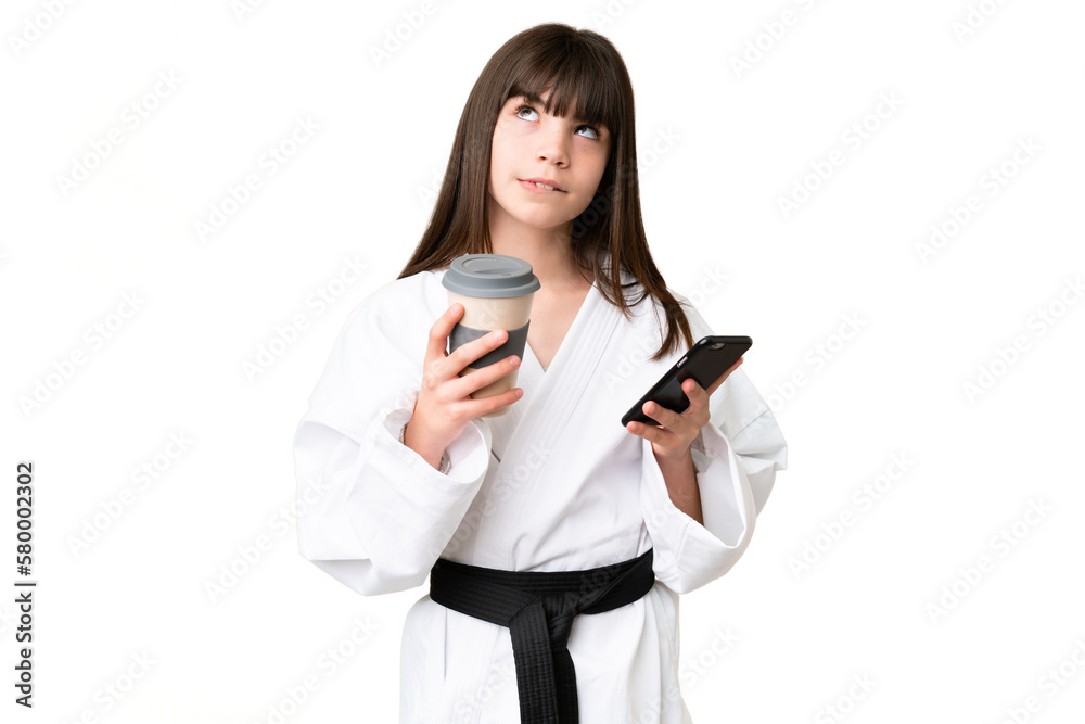 Little Caucasian girl doing karate over isolated background holding coffee to take away and a mobile while thinking something Little Caucasian girl doing karate over isolated background holding coffee to take away and a mobile while thinking something