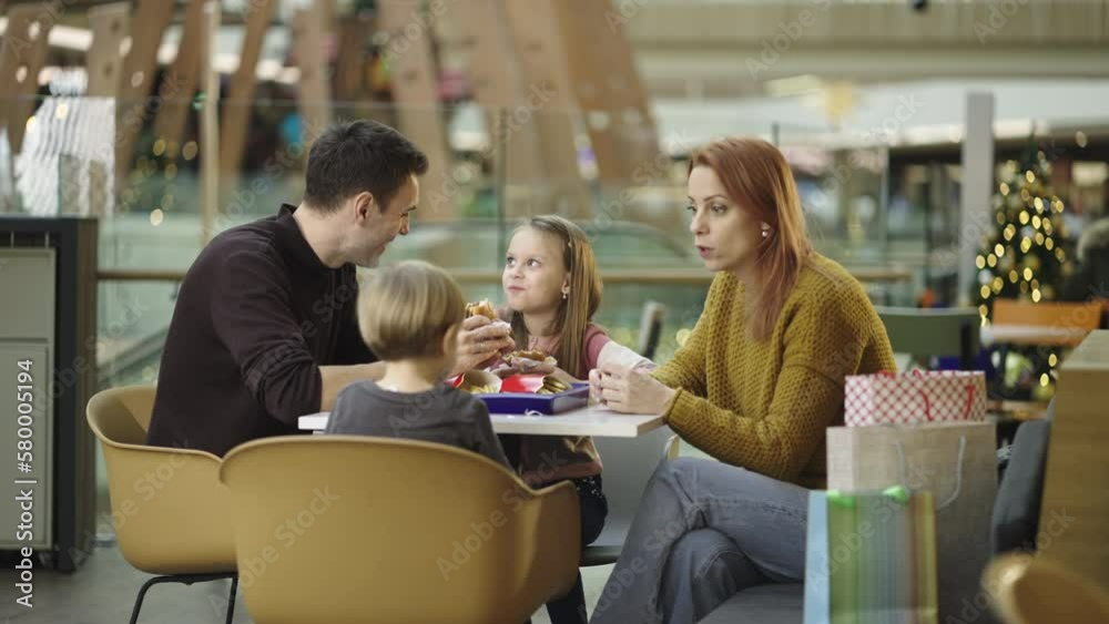 Beautiful family with two kids enjoying fast food after shopping in a modern restaurant. Father having fun with kids while eating burger in slow-motion Beautiful family with two kids enjoying fast food after shopping in a modern restaurant. Father having fun with kids while eating burger in slow-motion