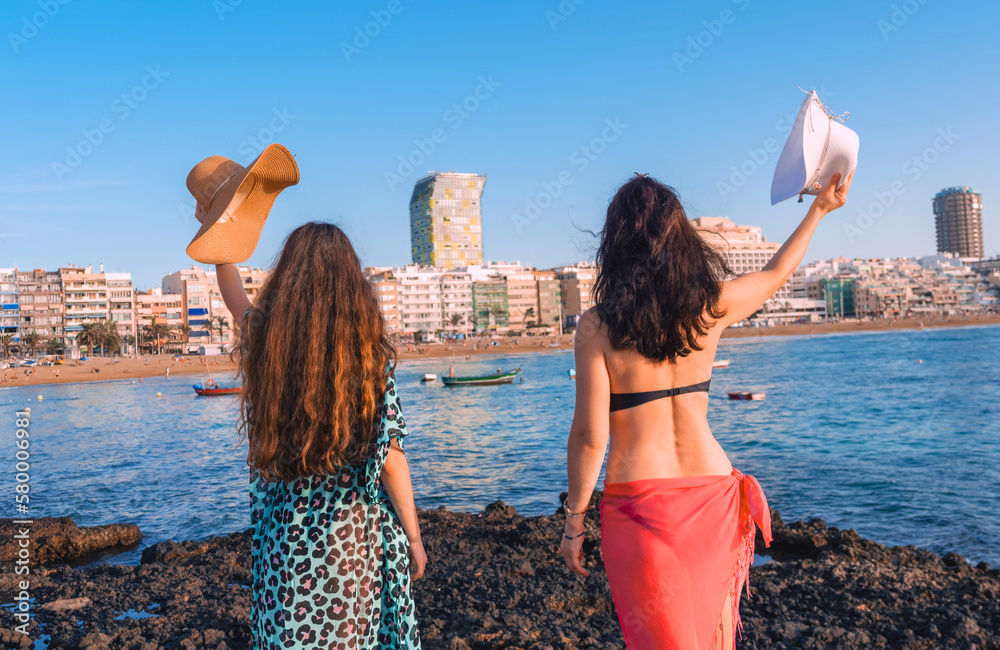 Rear view of two beautiful women on summer vacation greeting the beach of Las Canteras raising their sun hats