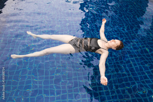 Woman lies on her back on the surface of the water of the pool, arms and legs spread out to the sides. Middle-aged woman swims in the pool. Woman in a black swimsuit relaxes in the pool.