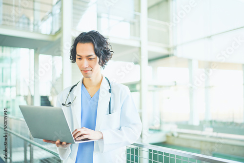 病院の廊下でノートパソコンを開くスクラブ白衣を着た若い男性医師