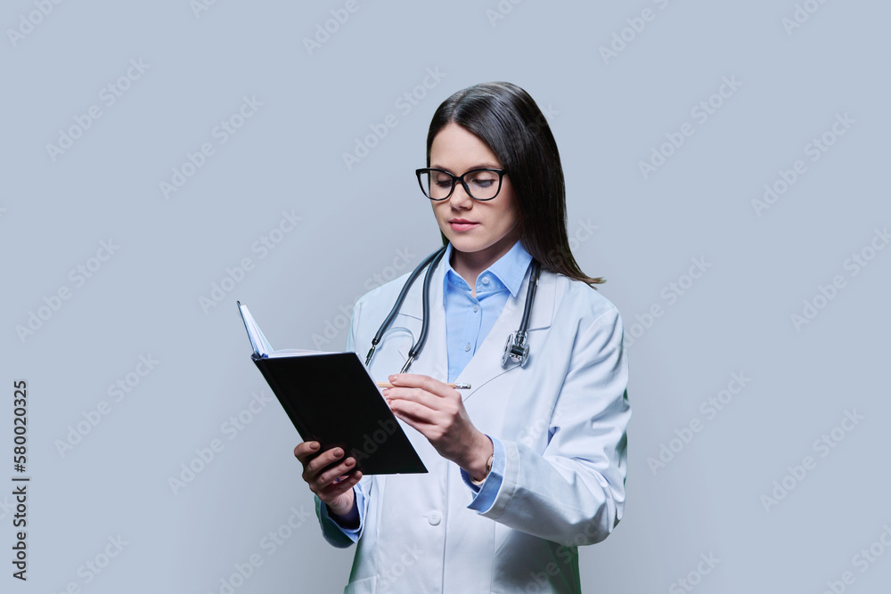 Young female doctor writes in notebook, on gray studio background. Young female doctor writes in notebook, on gray studio background.