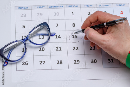 Kartka z kalendarza i dłoń z długopisem zaznaczająca datę obok leżą okulary