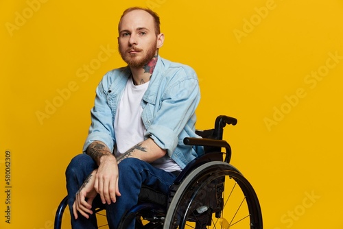 A man in a wheelchair looks at the camera, with tattoos on his arms sits on a yellow studio background, the concept of health is a person with disabilities