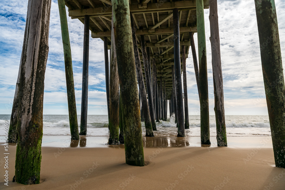 The view under the fishing pier in Ocean City, Maryland.  In the background waves gently roll in under cloudy blue skies. 