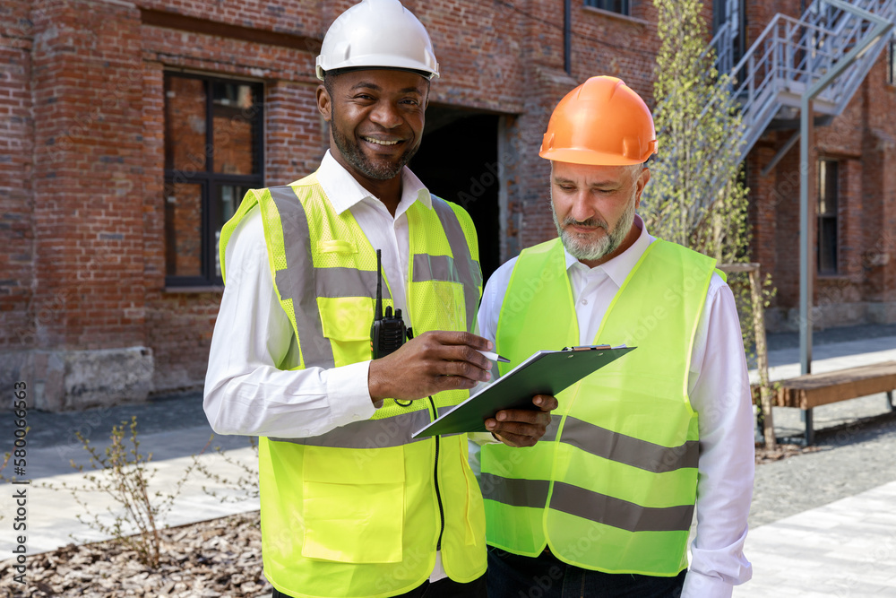 Portrait of the african american architect with blueprint sharing reconstruction ideas while senior caucasian colleague looking on notes. Coworkers standing near construction wall on fresh air. Portrait of the african american architect with blueprint sharing reconstruction ideas while senior caucasian colleague looking on notes. Coworkers standing near construction wall on fresh air.