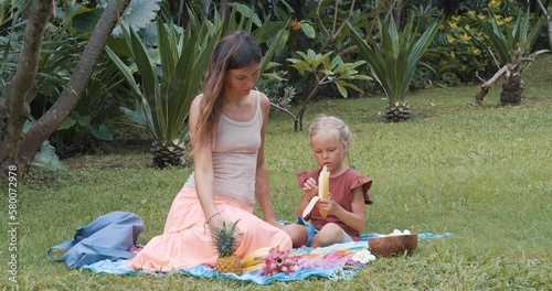Mom and daughter spending time together eating fruits sitting on the grass. Picnic, motherhood and weekend concept