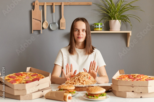 Portrait of serious confident woman with brown hair wearing white casual T-shirt sitting at table in kitchen  keeps diet  showing stop gesture  doesn t eat junk food  prefers healthy eating.