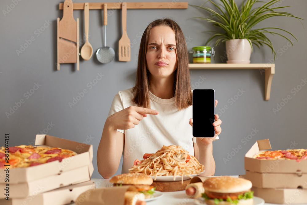 Portrait of sad winsome Caucasian woman wearing white casual T-shirt sitting at table in kitchen, being surrounded with junk food, pointing cell phone with empty screen, empty display.