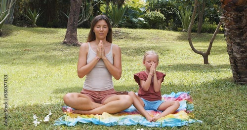 Young mother with her daughter meditating with closed eyes sitting in the lotus position on the grass in the park.