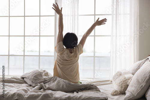 Satisfied relaxed middle aged lady sitting on bed, rising hands, looking at large window, enjoying good morning, feeling full of energy after healthy sleeping on comfortable mattress