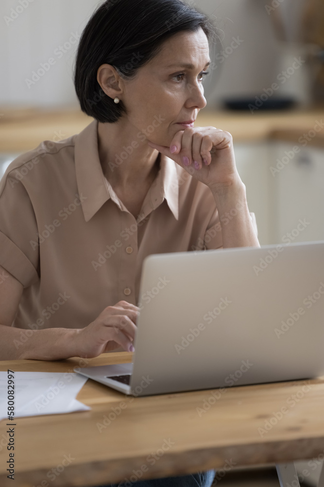 Serious pensive mature freelancer lady working at laptop from home, sitting at kitchen table, looking away with thoughtful face, touching chin, thinking, making decision in deep thoughts Serious pensive mature freelancer lady working at laptop from home, sitting at kitchen table, looking away with thoughtful face, touching chin, thinking, making decision in deep thoughts