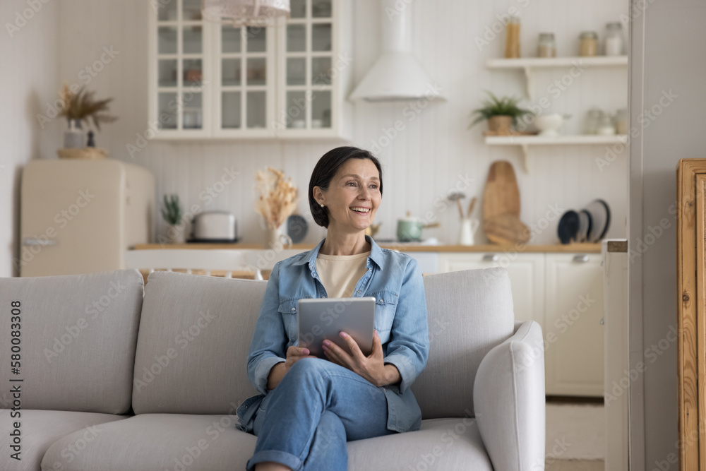 Cheerful pensive mature business owner woman sitting on couch, using tablet computer for online communication, looking away, smiling, enjoying work from home, thinking over freelance job success