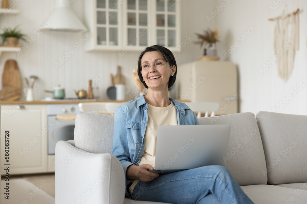 Happy mature freelancer woman holding laptop computer on lap, sitting on soft couch, looking away with positive thoughts, smiling, enjoying online communication, thinking, dreaming Happy mature freelancer woman holding laptop computer on lap, sitting on soft couch, looking away with positive thoughts, smiling, enjoying online communication, thinking, dreaming