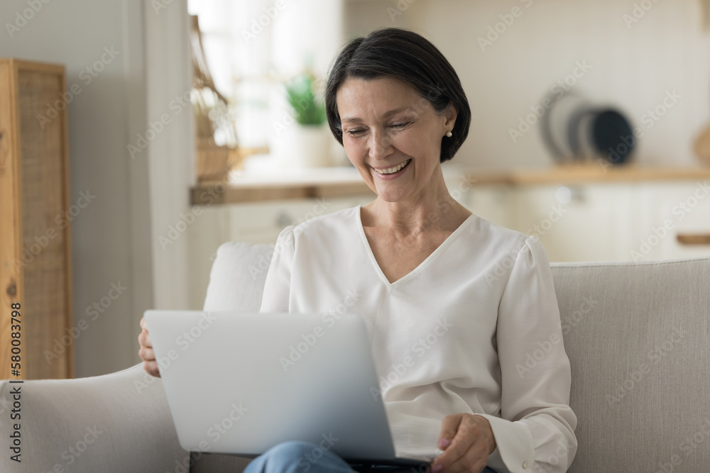 Cheerful pretty senior retired business woman using laptop at home, resting on comfortable sofa, talking on video call, looking at screen, smiling, laughing, enjoying online communication Cheerful pretty senior retired business woman using laptop at home, resting on comfortable sofa, talking on video call, looking at screen, smiling, laughing, enjoying online communication