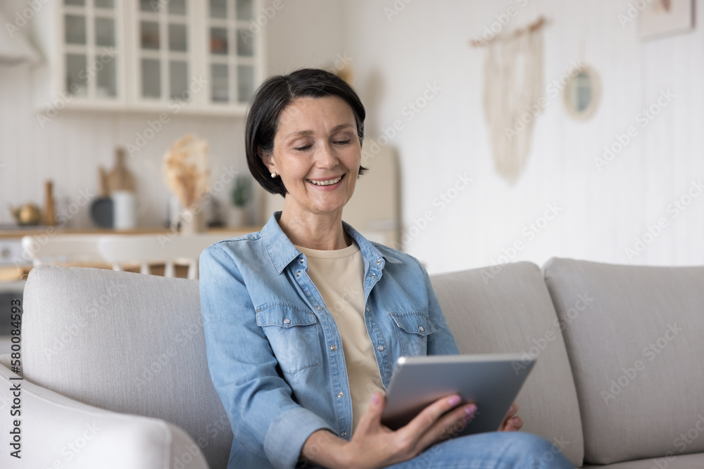 Happy relaxed senior lady watching media content, movie on tablet computer, sitting on sofa at home, looking at screen, smiling, laughing, making video call, enjoying conference talk Happy relaxed senior lady watching media content, movie on tablet computer, sitting on sofa at home, looking at screen, smiling, laughing, making video call, enjoying conference talk