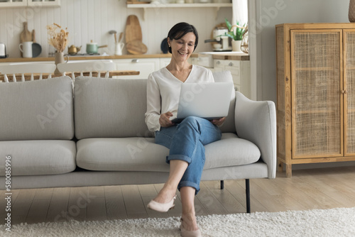 Happy attractive mature freelancer woman working at laptop at home, holding computer on lap, sitting on couch, talking on video call, looking at screen, laughing, enjoying Internet communication