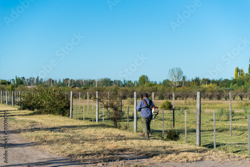 A man walks along a fence with a blue bag on his shoulder.