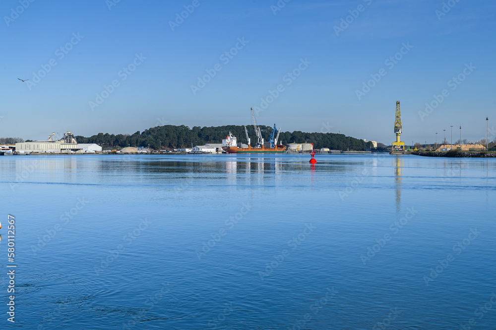 port of bayonne, france, in the background a red merchandise ship moored to the pier, in the foreground the water of the bay is calm, calm, above the blue sky, without clouds, of a sunny winter mornin