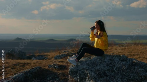 Young woman listening to music in headphones while sitting on a stone outdoors on a mountain with an incredible view, dancing, singing along while enjoying her favorite song while walking or traveling