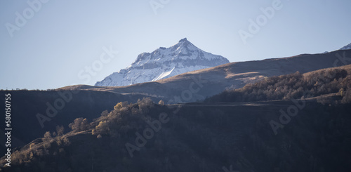 Panorama of mountain slopes with vegetation and trees yellowed in autumn and a rocky massif with snow, early autumn morning