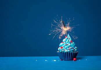 Blue cupcake with red and white sprinkles and lit sparkler on a blue background.