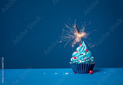 Blue cupcake with red and white sprinkles and lit sparkler on a blue background.